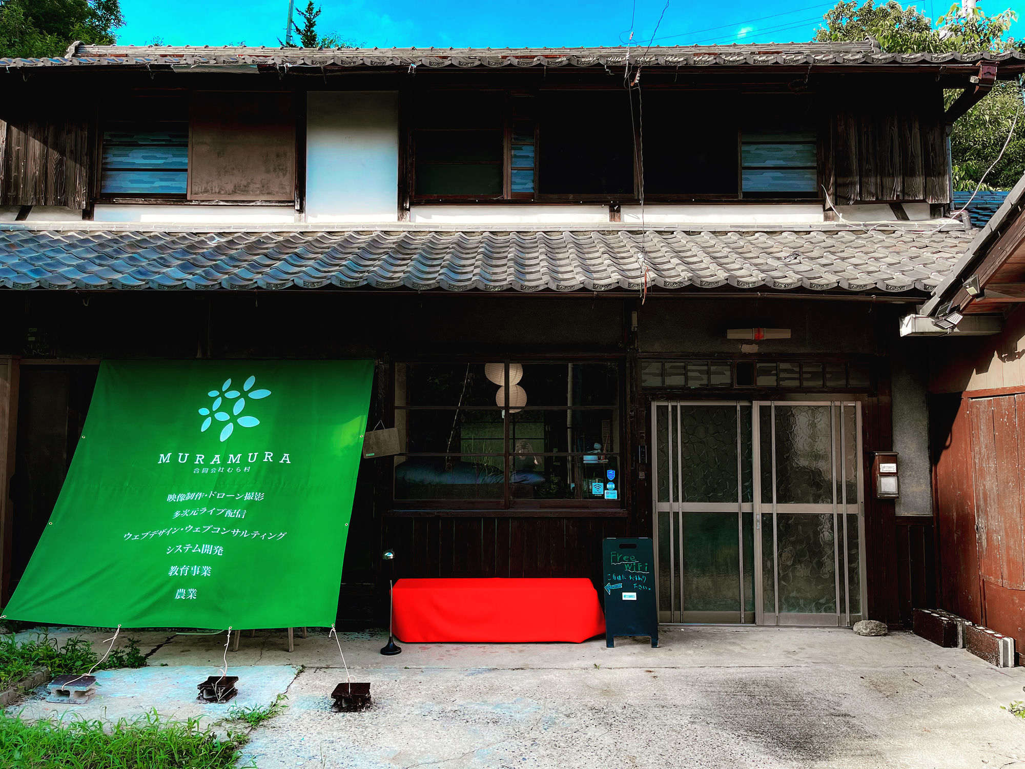 京都府南山城村にある MURAMURA の事務所外観 — 瓦屋根の古民家に事業内容を記した緑の垂れ幕と Free Wi-Fi の看板